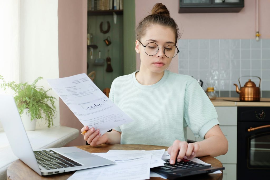 A woman wearing glasses sits at a table with a laptop and papers, focused on her work.